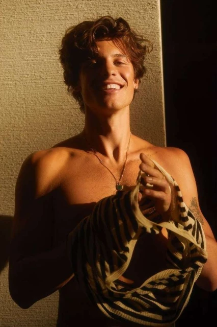 Shirtless Shawn Mendes smiles in a close-up studio shot. He holds a striped top with a necklace. Warm light highlights his features.