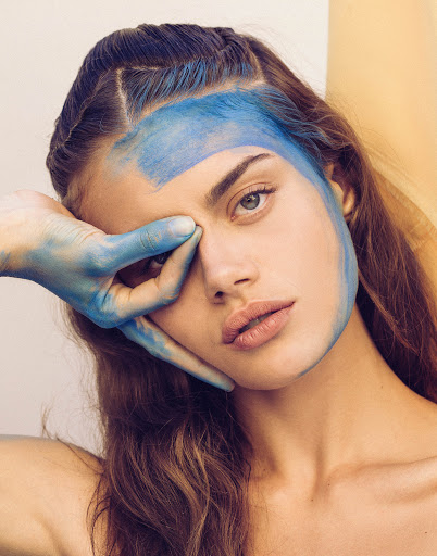 Close-up portrait of a woman with blue paint on her face and hand covering one eye, photographed by Sami Drasin.