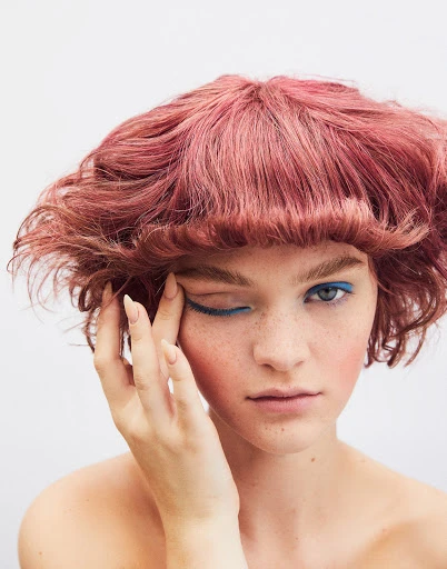 Close-up shot of a woman with pink hair, blue eye makeup, and fair skin posing in front of a bright white background.