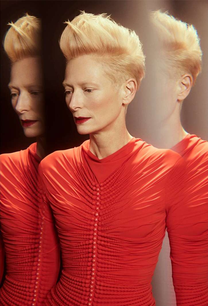 A striking portrait features a person with short, styled blonde hair and red lipstick, wearing a red, long-sleeved dress with gathered details.