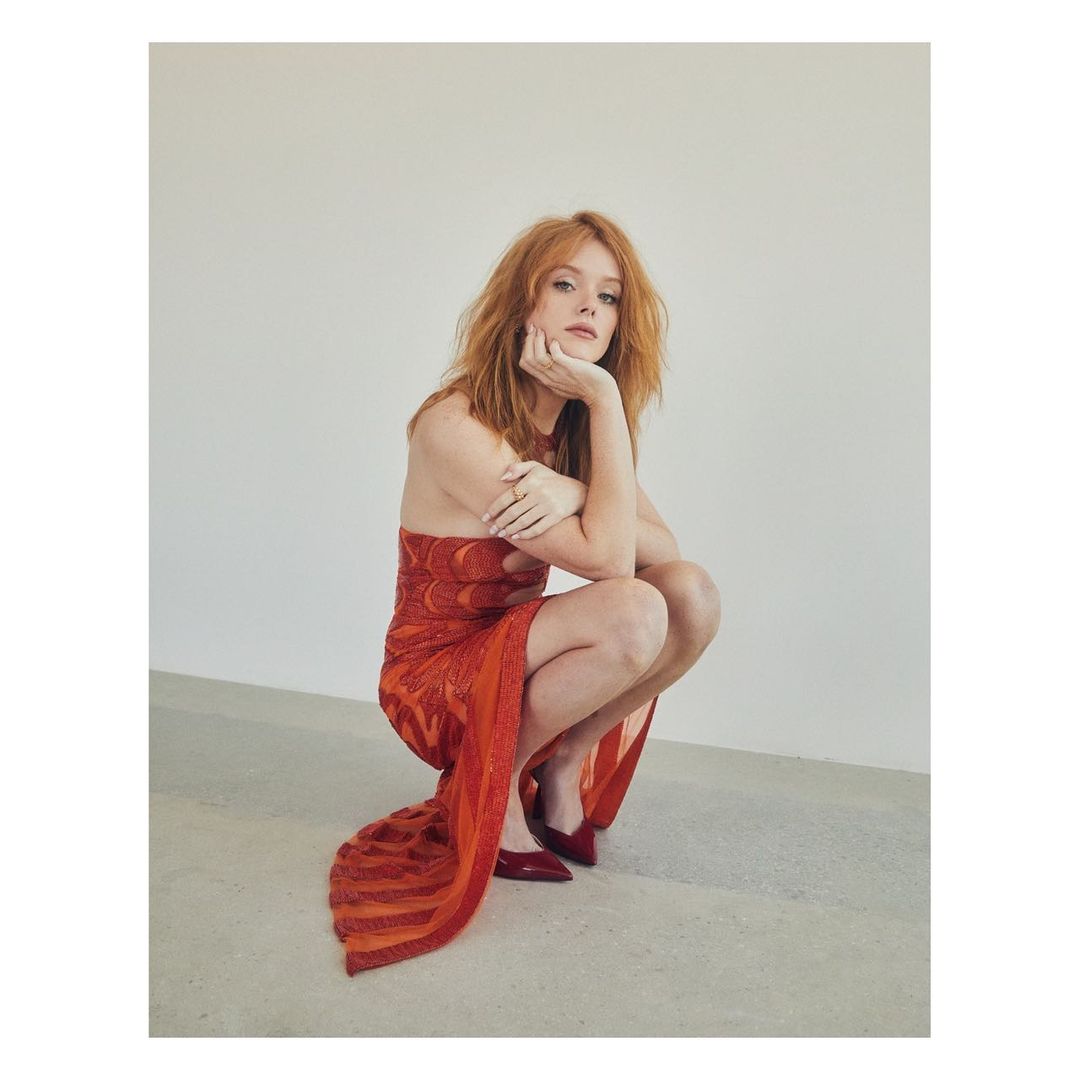 Red-haired woman in an orange dress crouching indoors, leaning her face on her hand, wearing red shoes against a neutral background.