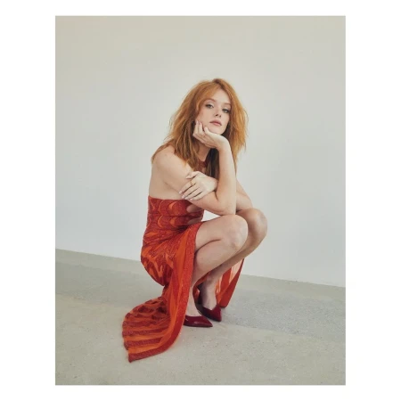 Red-haired woman in an orange dress crouching indoors, leaning her face on her hand, wearing red shoes against a neutral background.