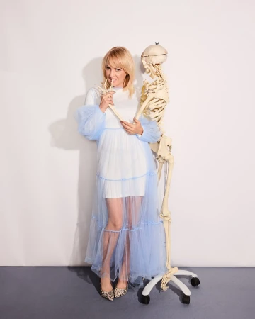 Blonde woman in dress poses with a human skeleton against a white background, holding a bone-like object, creating an edgy yet playful image.