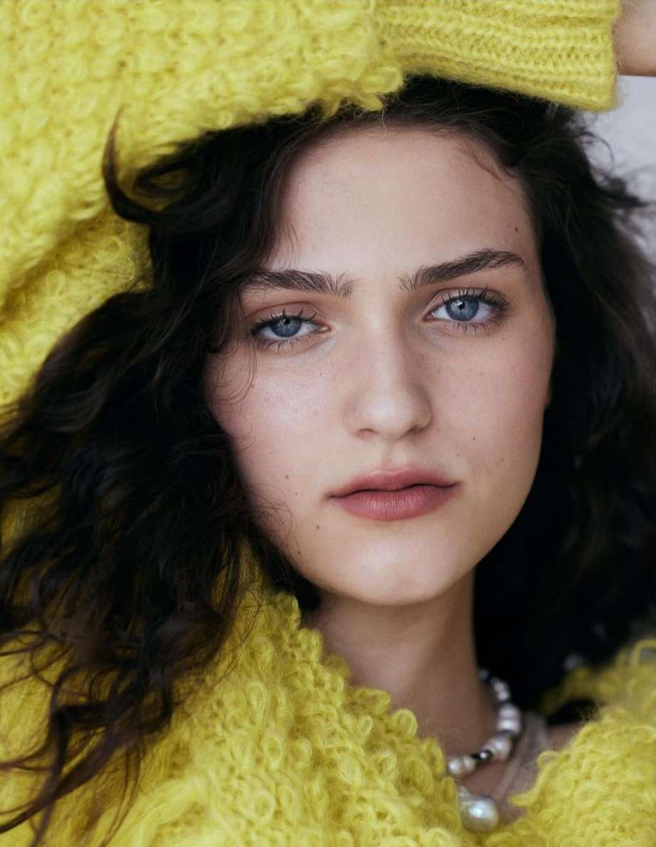 Close-up portrait of a woman with dark curly hair wearing a textured yellow sweater and pearl necklace