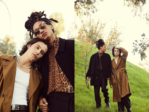 Two adult males in fashionable coats pose outside against a greenery backdrop in natural light, highlighting contemporary fashion apparel.