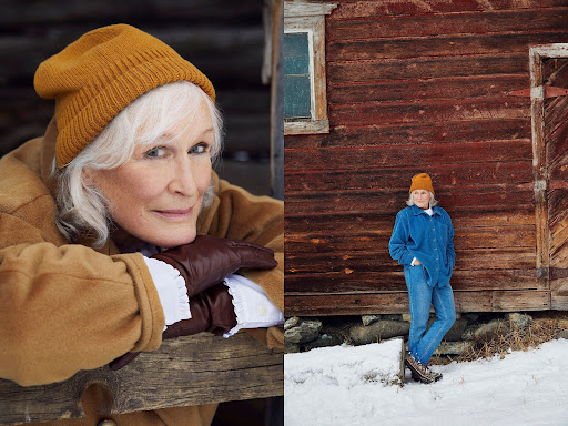 Glenn Close poses in a warm hat and jacket against a rustic wooden backdrop; standing outside a barn in winter snow, wearing a blue denim outfit.