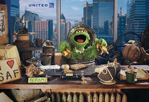 Image of Oscar the Grouch in a trash can on a desk with a window view of city skyscrapers and the United Airlines logo in the corner.