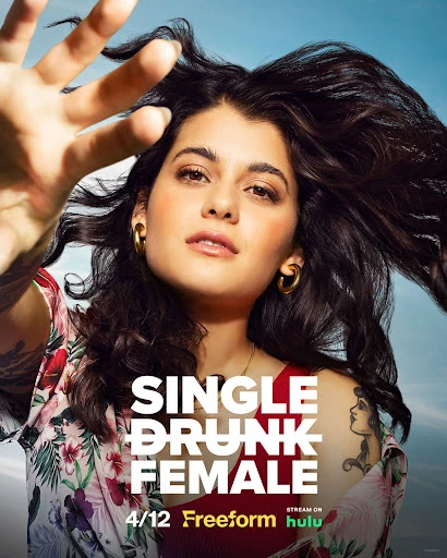 Portrait of woman with dark hair wearing earrings, posing with hand outstretched against a light blue background for TV show Single Drunk Female.