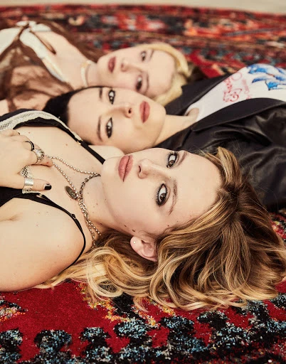 Eye-level group portrait of three young adult women with fair skin and a mix of blonde and brunette hair, lying on an ornate carpet.