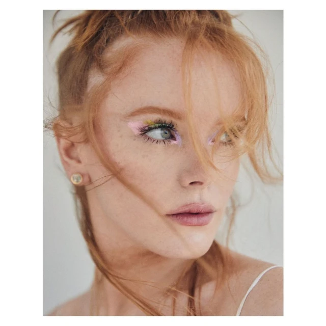 Close-up portrait of a redhead woman with green and pink eyeshadow looking to the right against a white background in a photo studio.