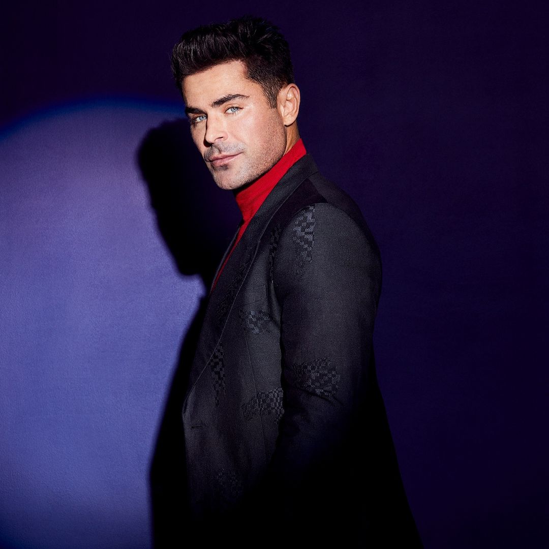 Zac Efron posing in a dark patterned suit and red turtleneck against a graduated purple background in a studio shot.