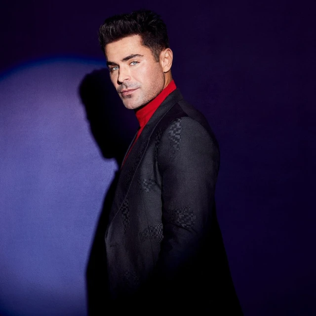 Zac Efron posing in a dark patterned suit and red turtleneck against a graduated purple background in a studio shot.