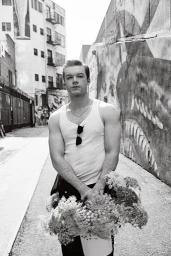 Portrait of Cameron Monaghan in a white tank top holding a large bouquet in an alley, photographed by Sami Drasin.