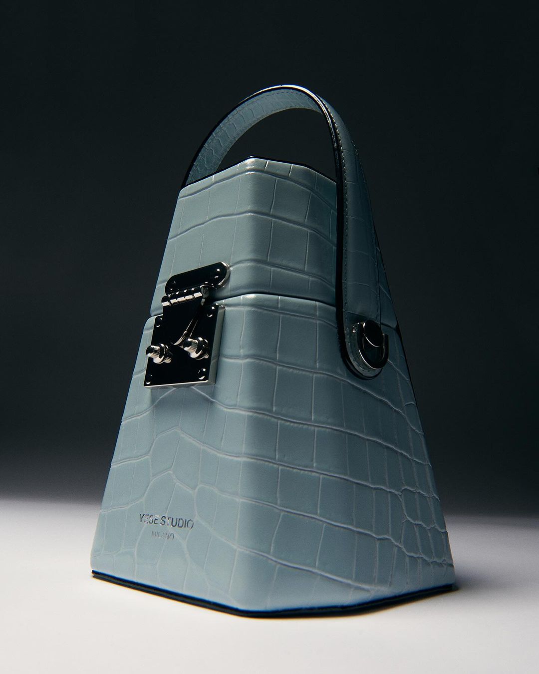 Trapezoid-shaped handbag with a handle, made of faux crocodile leather in a light blue color, featuring silver hardware and a logo.