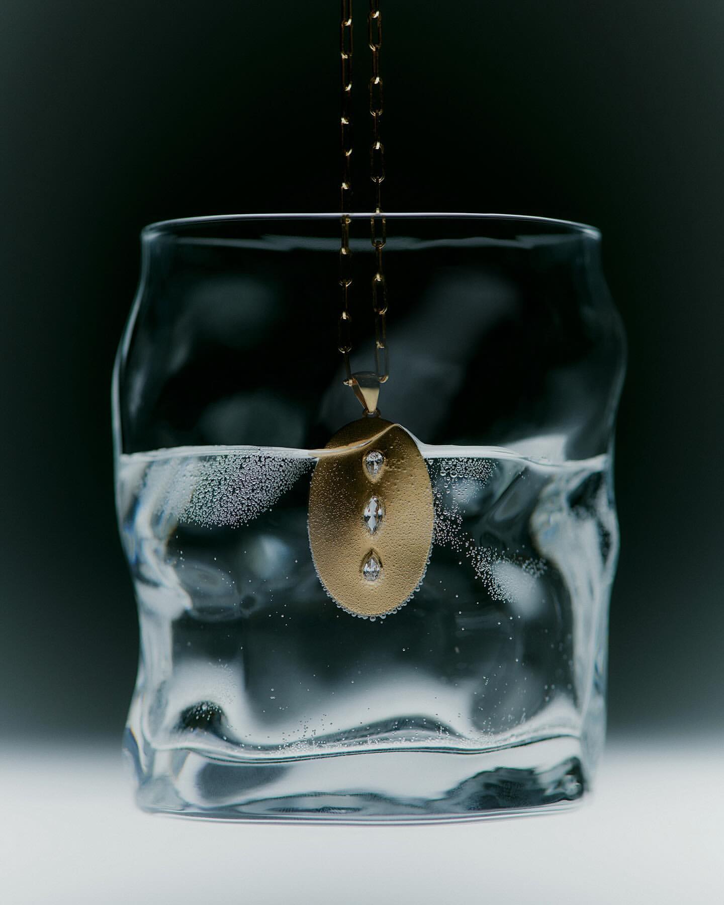 Gold pendant necklace with diamonds submerged in a textured drinking glass of clear water on a dark, cool-toned gradient background.