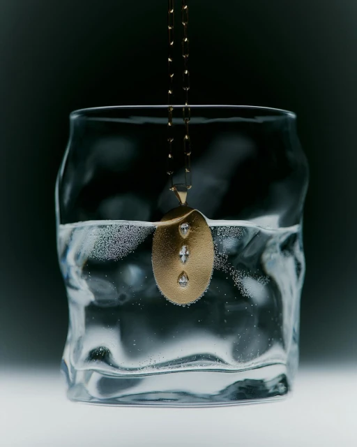 Gold pendant necklace with diamonds submerged in a textured drinking glass of clear water on a dark, cool-toned gradient background.