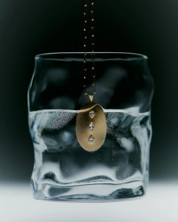 Gold pendant necklace with diamonds submerged in a textured drinking glass of clear water on a dark, cool-toned gradient background.
