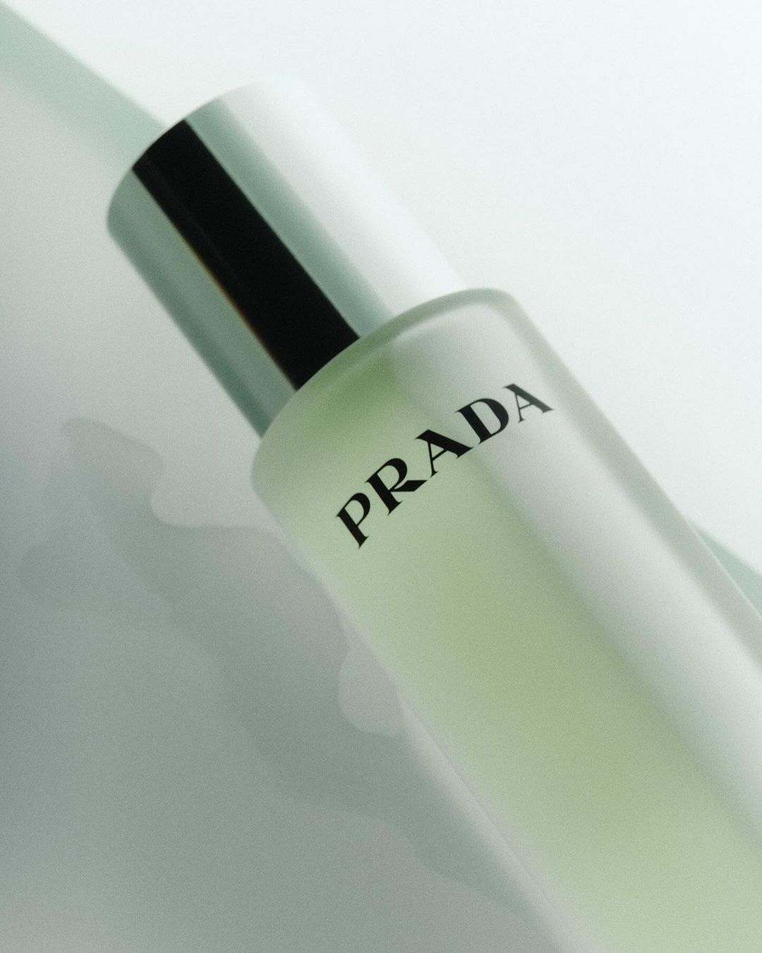 Close-up product shot of a Prada perfume bottle with a silver cap and black text on a light surface. Beauty and fashion advertising.