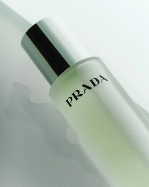 Close-up product shot of a Prada perfume bottle with a silver cap and black text on a light surface. Beauty and fashion advertising.