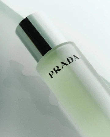 Close-up product shot of a Prada perfume bottle with a silver cap and black text on a light surface. Beauty and fashion advertising.