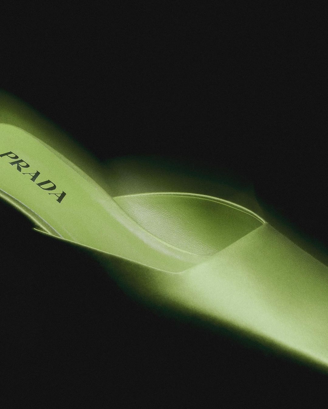 Close-up studio shot of a single, bright green, satin stiletto heel with the Prada logo on the insole against a dark background.
