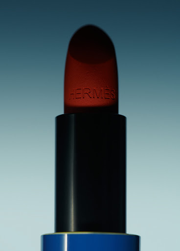 Close-up studio shot of a red Hermès lipstick with a black tube and blue base against a soft light blue background.