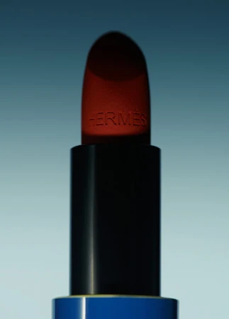 Close-up studio shot of a red Hermès lipstick with a black tube and blue base against a soft light blue background.