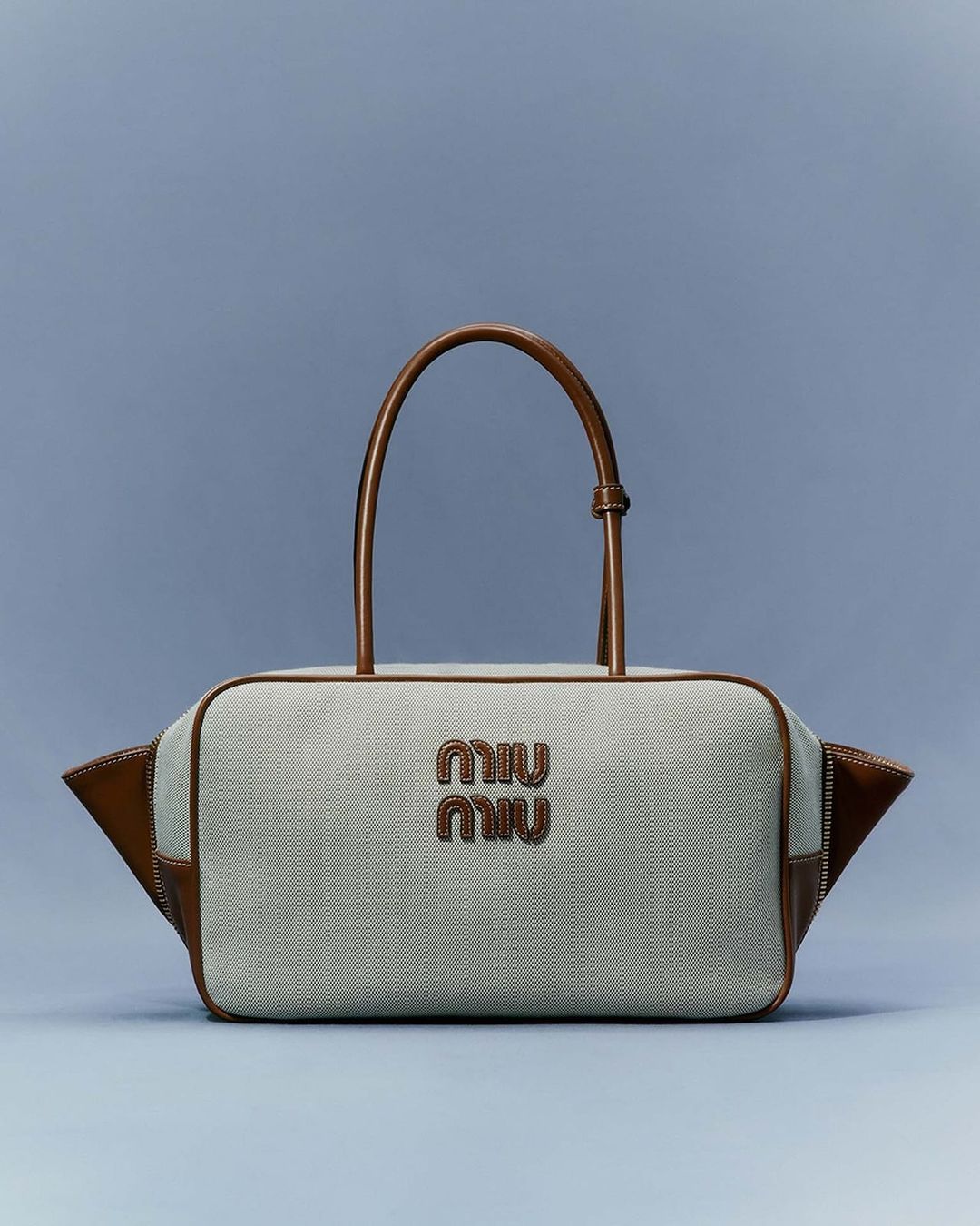 Tan and grey fabric satchel handbag with leather trim and handles, featuring a designer logo against a plain blue background in studio lighting.