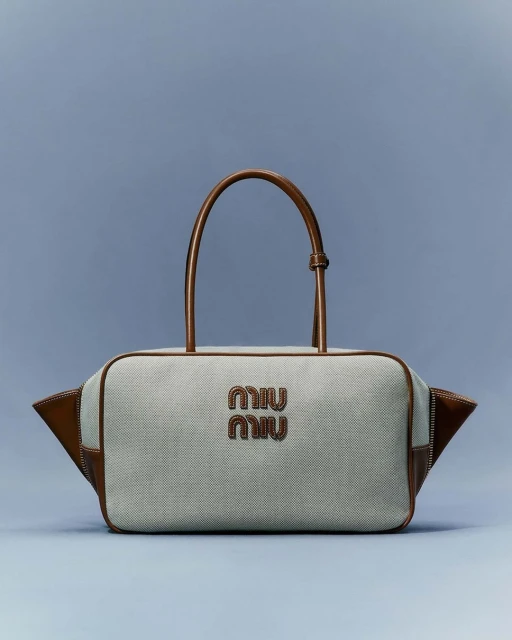 Tan and grey fabric satchel handbag with leather trim and handles, featuring a designer logo against a plain blue background in studio lighting.