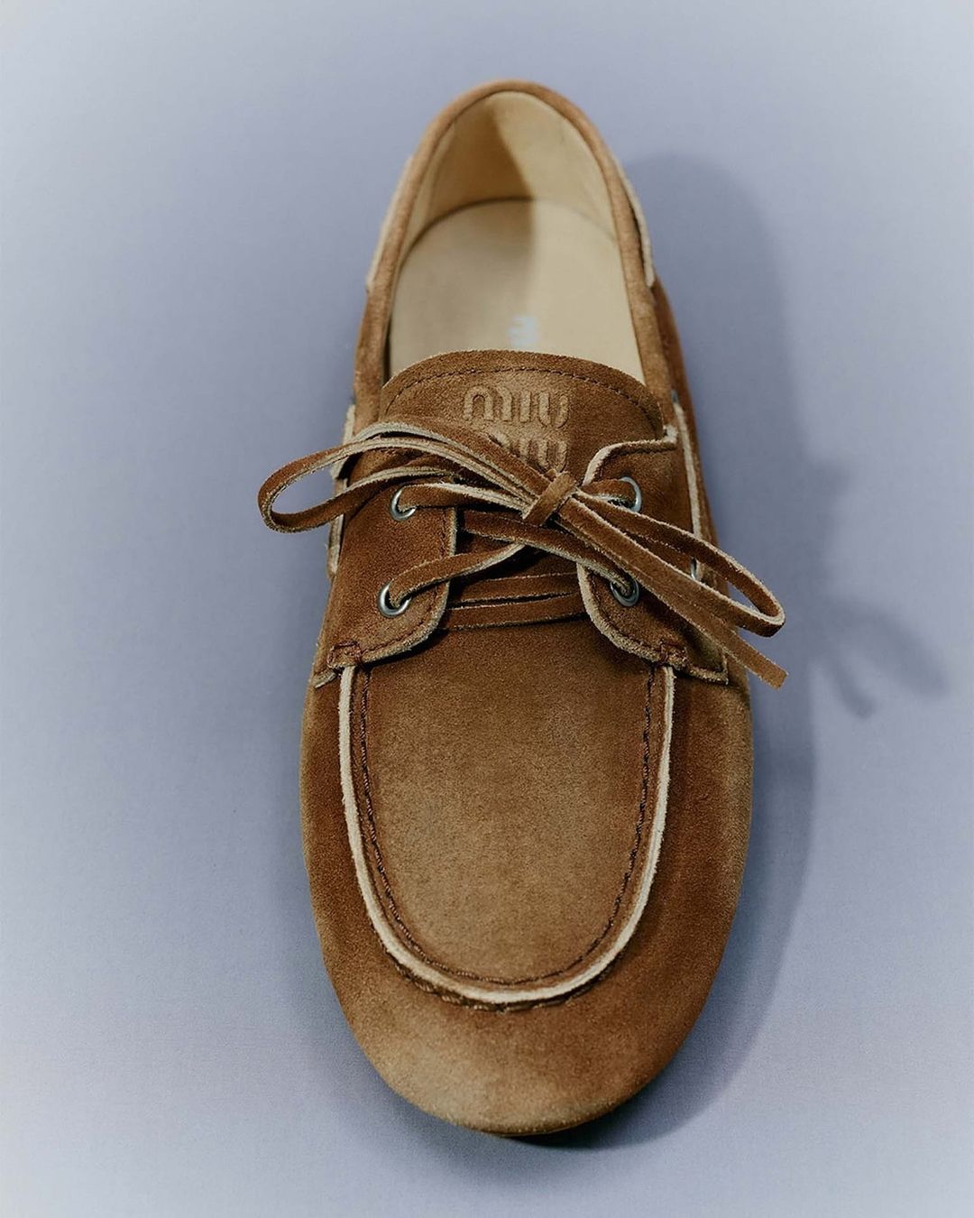 Studio shot of a brown suede boat shoe with white trim and laces, brand logo embroidered on the shoe, neutral gray background.