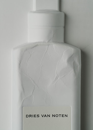Close-up studio shot of a white Dries Van Noten perfume bottle against a white backdrop with visible paper wrapping texture.