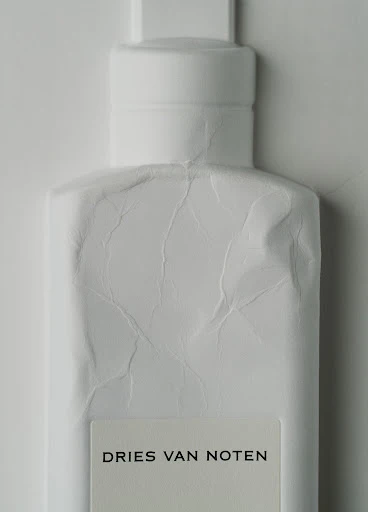 Close-up studio shot of a white Dries Van Noten perfume bottle against a white backdrop with visible paper wrapping texture.