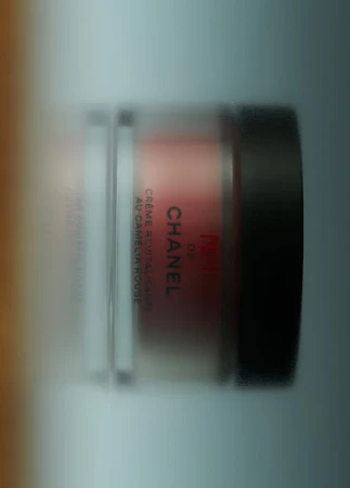 Close-up of a Chanel cream jar in a clear container with a black lid. The label is visible, showcasing brand text.