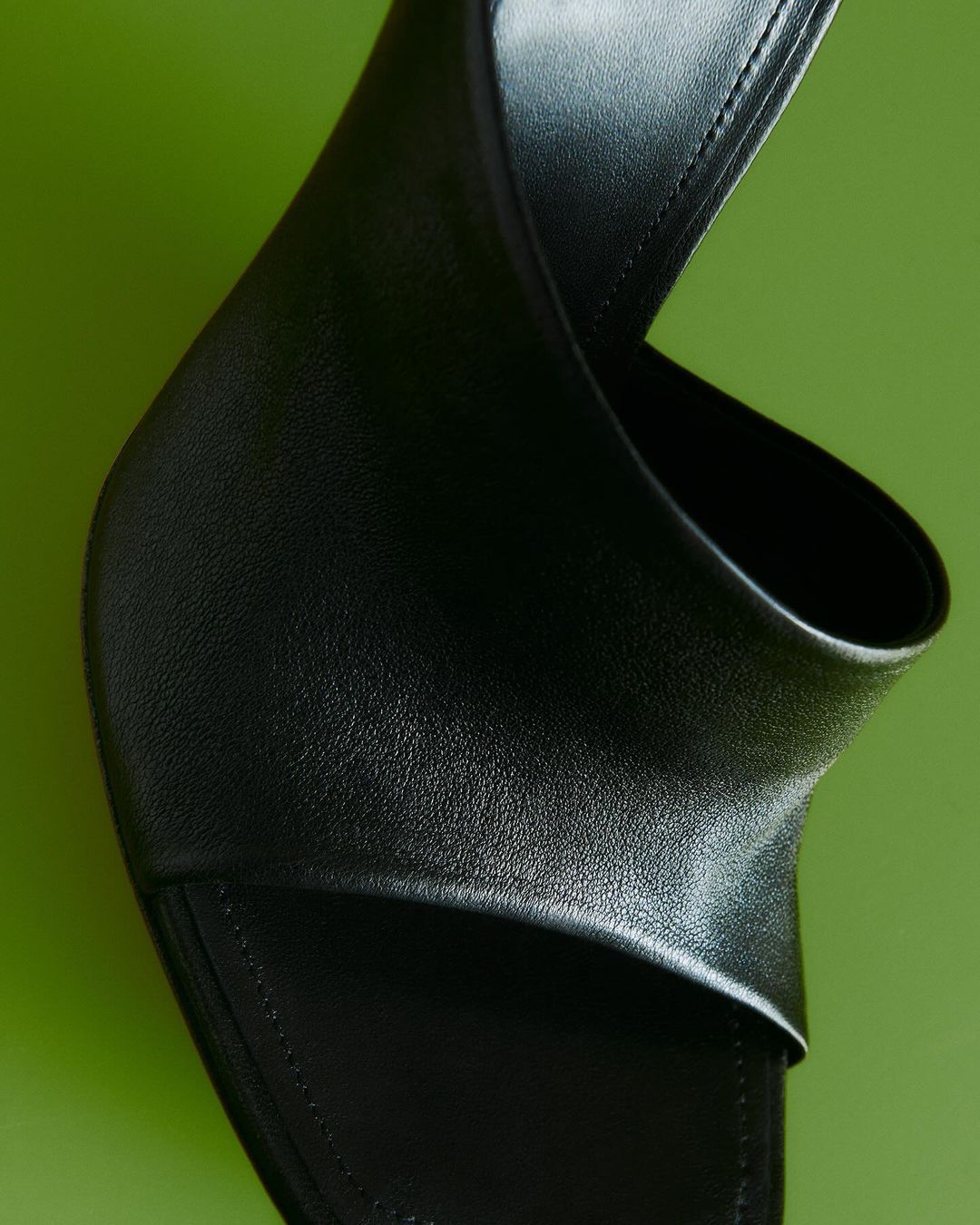 Close-up studio shot of a modern, open-toe black leather mule sandal, highlighting its design and texture against a green backdrop.