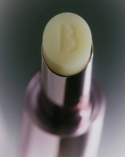Close-up of a golden-pink lip balm tube with a pale yellow balm top featuring an embossed 'B' logo, set against a soft grey background.