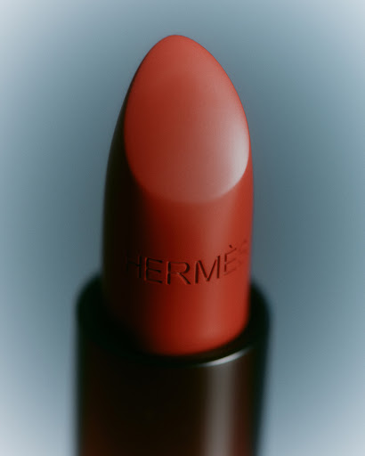 Close-up of a red-orange lipstick tube with 'Hermès' engraved on the side, set against a soft, graduated blue background.