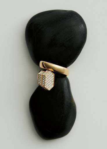 Still life of a gold ring and pendant jewelry piece balancing on two smooth black stones against a light neutral background.