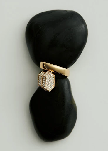 Still life of a gold ring and pendant jewelry piece balancing on two smooth black stones against a light neutral background.