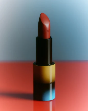 High-angle product shot of Hermès lipstick in a colourful tube, set against a graduated blue and red backdrop with soft lighting.