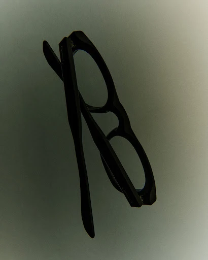 Black eyeglasses with a classic frame design are angled slightly on a neutral gray background.