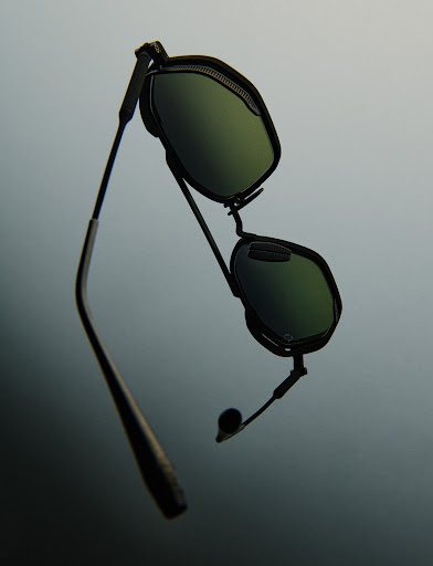 Floating black sunglasses with green lenses, featuring a sleek design and dark metallic frames against a blurred grey background.