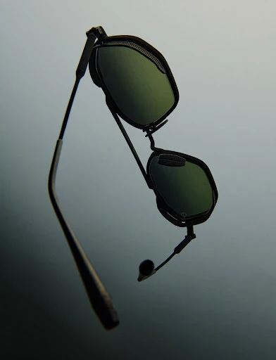 Floating black sunglasses with green lenses, featuring a sleek design and dark metallic frames against a blurred grey background.