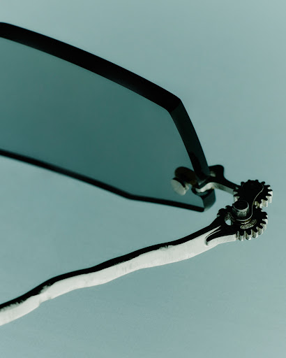 Macro shot of dark tinted sunglasses with metallic gear hinge details against a pale grey background