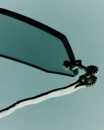 Macro shot of dark tinted sunglasses with metallic gear hinge details against a pale grey background