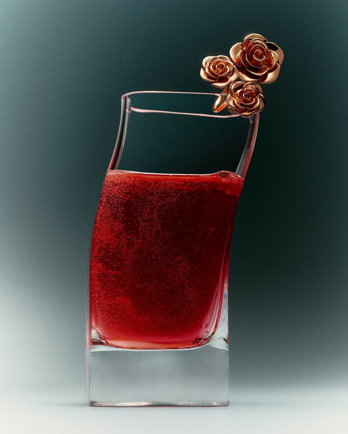 Close-up of a crystal glass filled with sparkling red liquid, adorned with three decorative golden rose embellishments on a muted background.