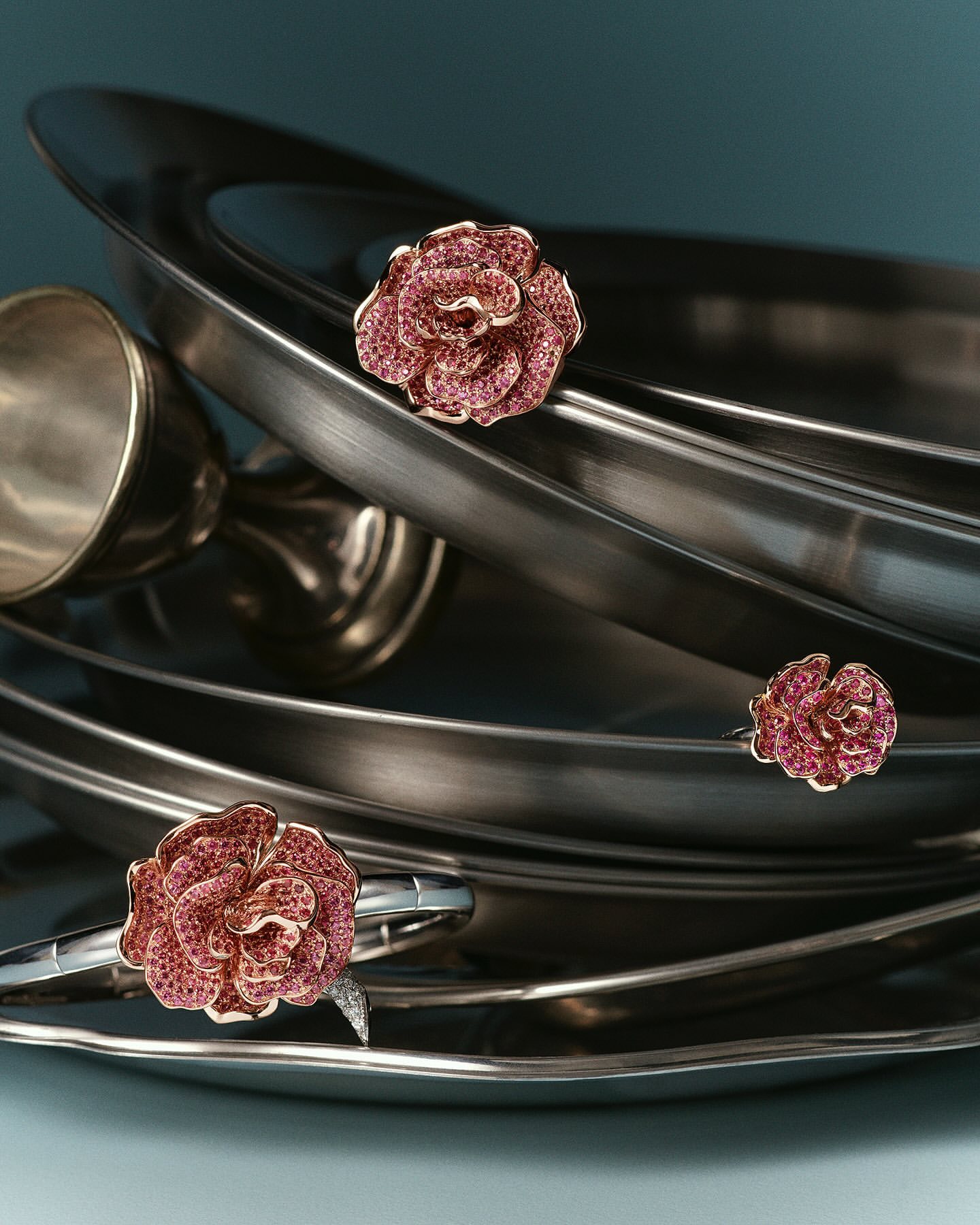 Close-up of a rose-shaped ring and a rose-shaped bracelet displayed on shiny, metal serving platters against a teal background.