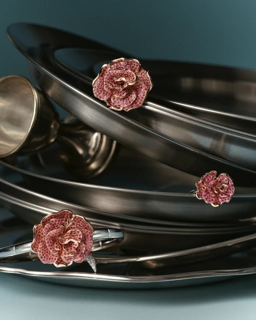 Close-up of a rose-shaped ring and a rose-shaped bracelet displayed on shiny, metal serving platters against a teal background.