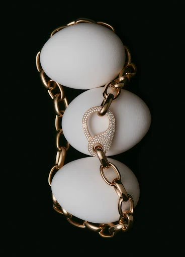 Close-up still life of a gold chain-link necklace draped over three white eggs against a dark backdrop, showcasing luxury jewelry.