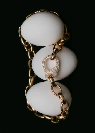 Close-up still life of a gold chain-link necklace draped over three white eggs against a dark backdrop, showcasing luxury jewelry.