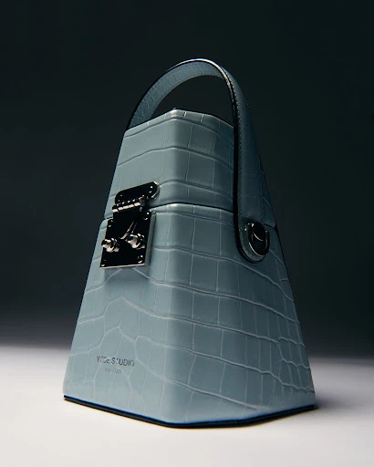 Light blue Yuzefi handbag featuring a mock crocodile texture, top handle, and silver hardware.  Studio product shot with gradient background.